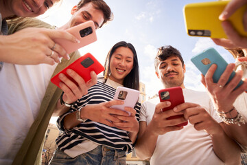 Low angle view multiracial group young generation z in circle using phones together outdoors. Cheerful student friends looking at mobile enjoying social media content. People and technology addiction  - Powered by Adobe