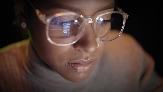 Close up of reflection of computer screen on the glasses of an African American hacker woman working on her laptop in the dark. Young people and security of computer networks and Internet. 