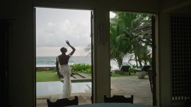 Flamboyant gay black male walks through luxury villa and poses by swimming pool in white elegant wedding dress. Gender fluid bride makes model photos on tropical island near palm trees and water.