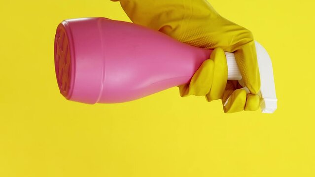 Vertical video. Housework tools. Hygiene product. Detergent equipment. Janitor shaking pink cleaner spray bottle in hand in protective glove isolated on yellow background.
