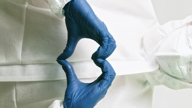 Vertical video. Medical care symbol. Love hands sign. Doctor in white lab protective coverall suit mask blue gloves with heart shape gesture.