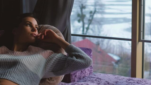 Lonely weekend. Idle leisure. Young beautiful pensive sad bored woman laying relaxing on couch during lazy pastime in cozy home interior with winter window view.