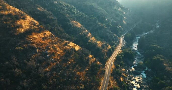 Rays of setting sun pass to the rocks of California desert. Lonely car runs by the wavy highway in mountains over the little quick river.