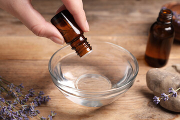 Woman dripping lavender essential oil from bottle into bowl at wooden table, closeup