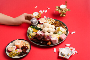 Female hand with diya lamps, candles and plates of different treats on red background. Divaly celebration