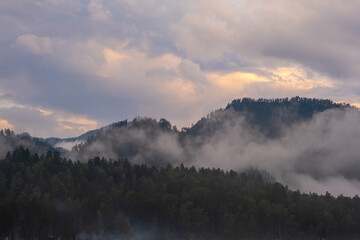 Fototapeta premium Mountains and peaks in fog at sunrise or sunset. Sun from behind the mountain. Amazing view of mountains and forest landscape with cloudy skies Altai mountains. Mountains and peaks in fog at sunrise