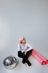A young woman with pink hair sits elegantly in a minimalist studio, wearing a white blouse, black...