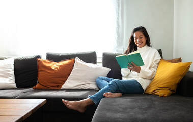 Happy, smiling Asian woman relaxing at home reading a book sitting on the sofa. Copy space.