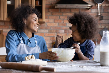 Fototapeta na wymiar Happy African American mother and little daughter baking at home together, kneading dough, smiling mom with 7s girl child preparing homemade pastry or cookies, enjoying leisure time in kitchen