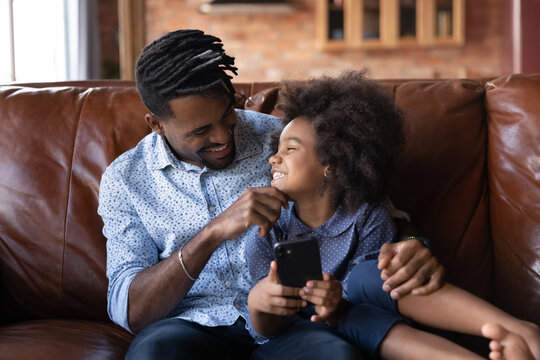 Happy African American father with little daughter using smartphone together, sitting on cozy couch at home, smiling dad and adorable 7s girl child having fun with phone, spending leisure time