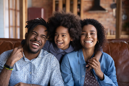 Head shot portrait happy African American parents with little daughter hugging, sitting on couch at home, smiling mother and father piggy backing adorable girl child looking at camera, family picture