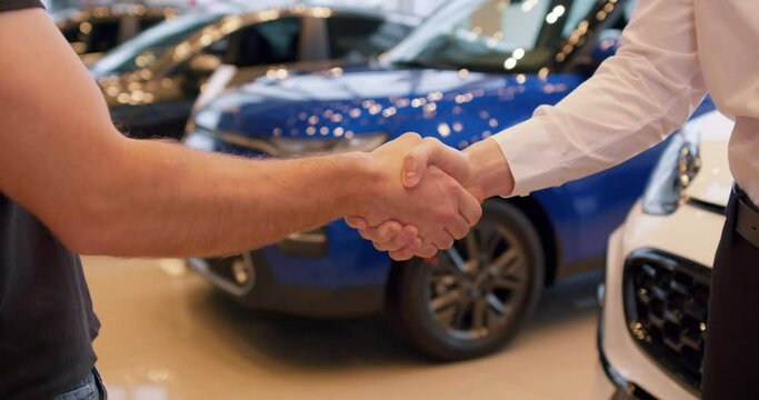 two men customer and client shaking hands in car dealership on background of bought cars. successful purchase, bargain in automotive dealership showroom. Slow motion, closeup cropped shot