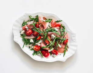 Delicious snack salad with prosciutto, arugula, cherry tomatoes, blue cheese on a light background, top view