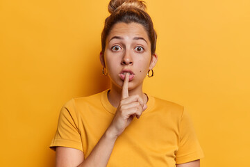 Fototapeta na wymiar Be silent please. Surprised young European woman keeps index finger over lips shares secret or asks to be quiet dressed in casual t shirt isolated over yellow background. Do not drop any word