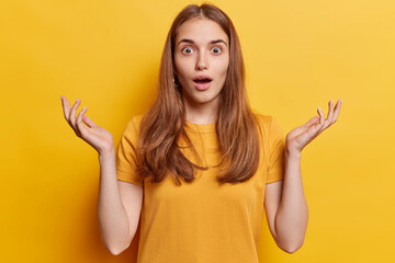 Surprised long haired young woman spreads palms and stares impressed at camera holds breath from amazement reacts to something shocking dressed in casual t shirt isolated over yellow background