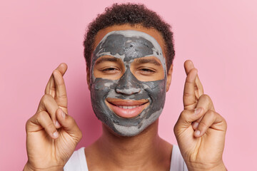Photo of pleased African man with dark curly hair applies nourishing clay mask on face keeps fingers crossed believes in good luck smiles happily isolated over pink background. Facial treatments