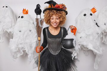 Cheerful curly haired woman wears hat and black dress holds broom and cauldron comes on Halloween...