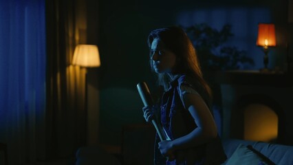 Young girl with scaried face expression walking in the dark living room, looking around, holding baseball bat in hand.