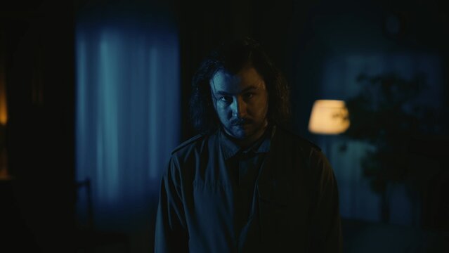 Man maniac with long hair standing in the dark apartment room, looking directly at the camera with straight face, tense atmosphere.