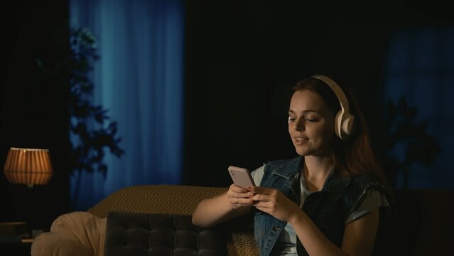 Girl in headphones sitting on the sofa in the dark living room, carefree listening to music on smartphone, smiling face expression.
