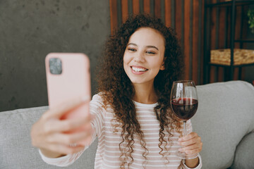 Young woman wear casual clothes sits on grey sofa couch do selfie shot on mobile cell phone drink...