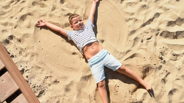 Happy child boy in a white striped T-shirt and blue summer shorts lies on the sand and makes an angel with his hands and feet