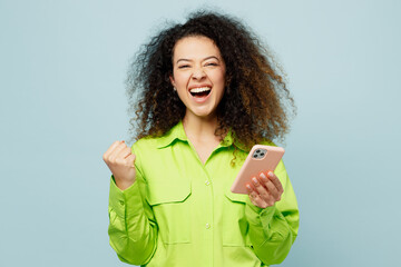 Young smiling happy latin woman she wears green shirt casual clothes hold in hand use mobile cell phone do winner gesture isolated on plain pastel light blue cyan background studio. Lifestyle concept. - obrazy, fototapety, plakaty