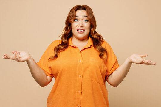 Young sad ginger chubby overweight woman wear orange shirt casual clothes spread hands shrugging shoulders look puzzled, have no idea isolated on plain pastel light beige background studio portrait