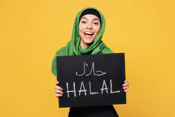 Young smiling arabian asian muslim woman wears green hijab abaya black clothes hold cardoard sign with halal text isolated on plain yellow background People uae middle eastern islam religious concept