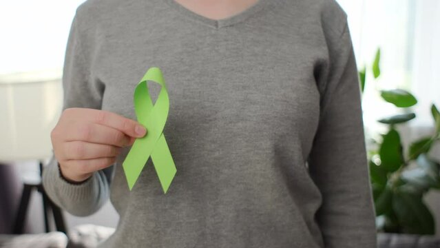Close-up of female hands holding small lime green ribbon standing in room. Achalasia, mental health, Non-Hodgkin lymphoma, Lyme disease awareness, world kidney day, organ donation concept
