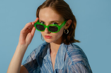 Fashionable young woman wearing trendy green sunglasses, chiffon blouse, posing on blue background....