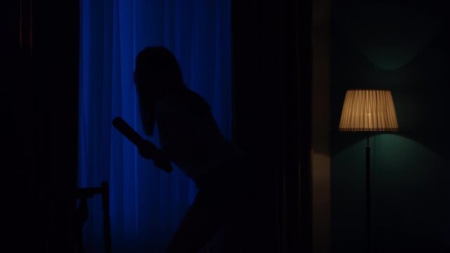 Closeup shot of afraid young girl with baseball bat standing near the window, looking around, thunderstorm outside, tense atmosphere. HDR BT2020 HLG Material.
