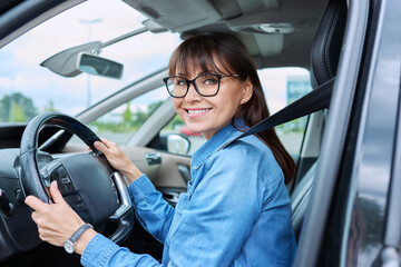 Middle-aged woman driver sitting behind wheel in car, smiling looking at camera