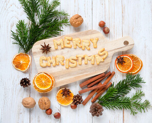 Christmas holiday background. cutting board with Merry Christmas text made of cookies, cinnamon, orange slices, anise, nuts, cones, fir branches on white wooden table. winter festive season. top view