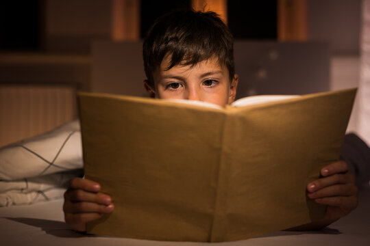 Bedtime story. Cute boy in the evening in his children's room lying on the bed holding an open book looking and reading fairy tale. Close-up of the book, only the boy's eyes are visible.