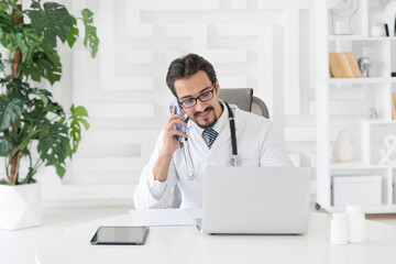 Close up portrait of smiling male doctor sitting at the desktop and talking to phone in the office...