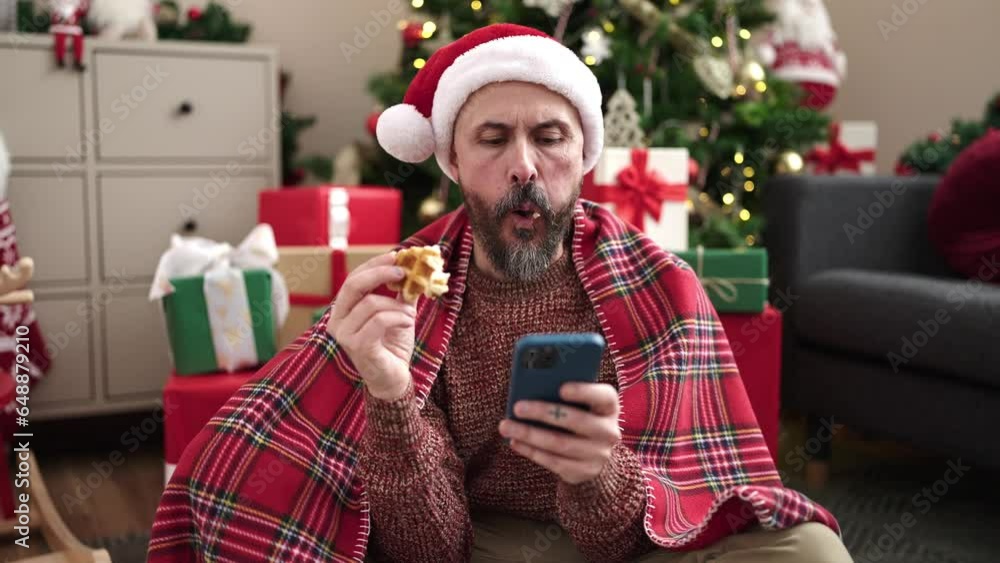 Sticker Young bald man using smartphone eating waffle sitting by christmas tree at home