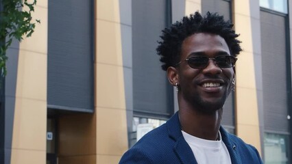 Fashionable african American male in stylish clothes stands in city next to modern building, looking with smile. Portrait Happy black man