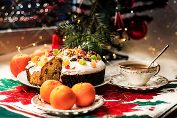 Obraz na płótnie Canvas Christmas stollen with glaze, candied fruits and nuts on a saucer with a cup of tea, tangerines near a small Christmas tree.