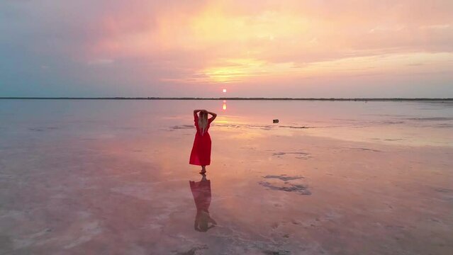 Aerial of a young woman in red dress standing in the water of a unique pink salt lake. Sunset at lake Bursol with beautiful reflections on calm water surface. Stunning scenery
