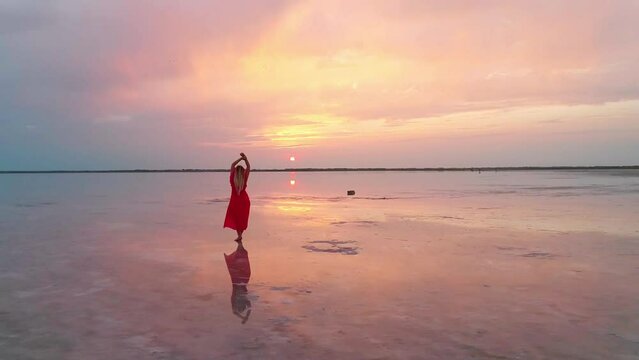 Aerial of a young woman in red dress standing in the water of a unique pink salt lake. Sunset at lake Bursol with beautiful reflections on calm water surface. Stunning scenery