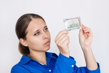 Young caucasian woman holding hundred-dollar cash banknote looking at the bill in the light checking it for authenticity isolated on a white background. Verification of the authenticity of money  - obrazy, fototapety, plakaty