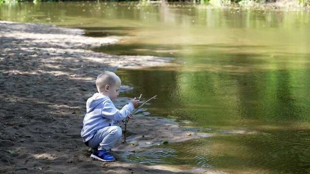 A kid boy traveler plays with sticks in the water on the river bank during a family hiking journey. A child plays on the sand on the river bank enjoying a family weekend.