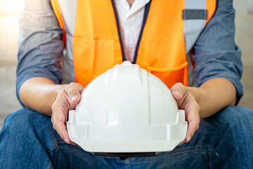 Male worker wearing reflective orange vest holding white protective safety helmet while sitting at...