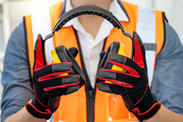 Male worker with reflective orange vest and protective hand gloves holding yellow safety ear muffs...