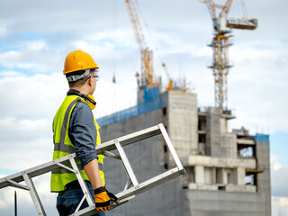 Asian man construction worker with green reflective vest, yellow safety helmet and ear muffs carrying aluminum ladder looking at unfinished building and tower crane at construction site © Summer Paradive