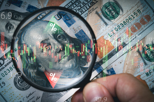 Hand holding magnifier glass to focus Benjamin Franklin face on USD dollar banknote with stock market chart for analysis investment on currency exchange or forex and economy inflation concept.