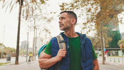 Mature man tourist wearing casual clothes, with backpack on his shoulder, looks around while standing on crosswalk