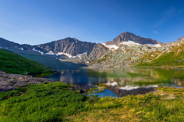 A serene mountain lake reflects the towering peaks and clear blue sky above, creating a tranquil and picturesque scene. The summer sun casts a warm glow, enhancing the beauty of the landscape.