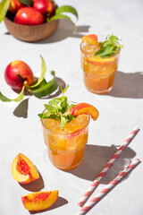 Two glasses with refreshing peach tea with ice and mint. Homemade cold summer drink on a light background with fresh fruits and shadows.